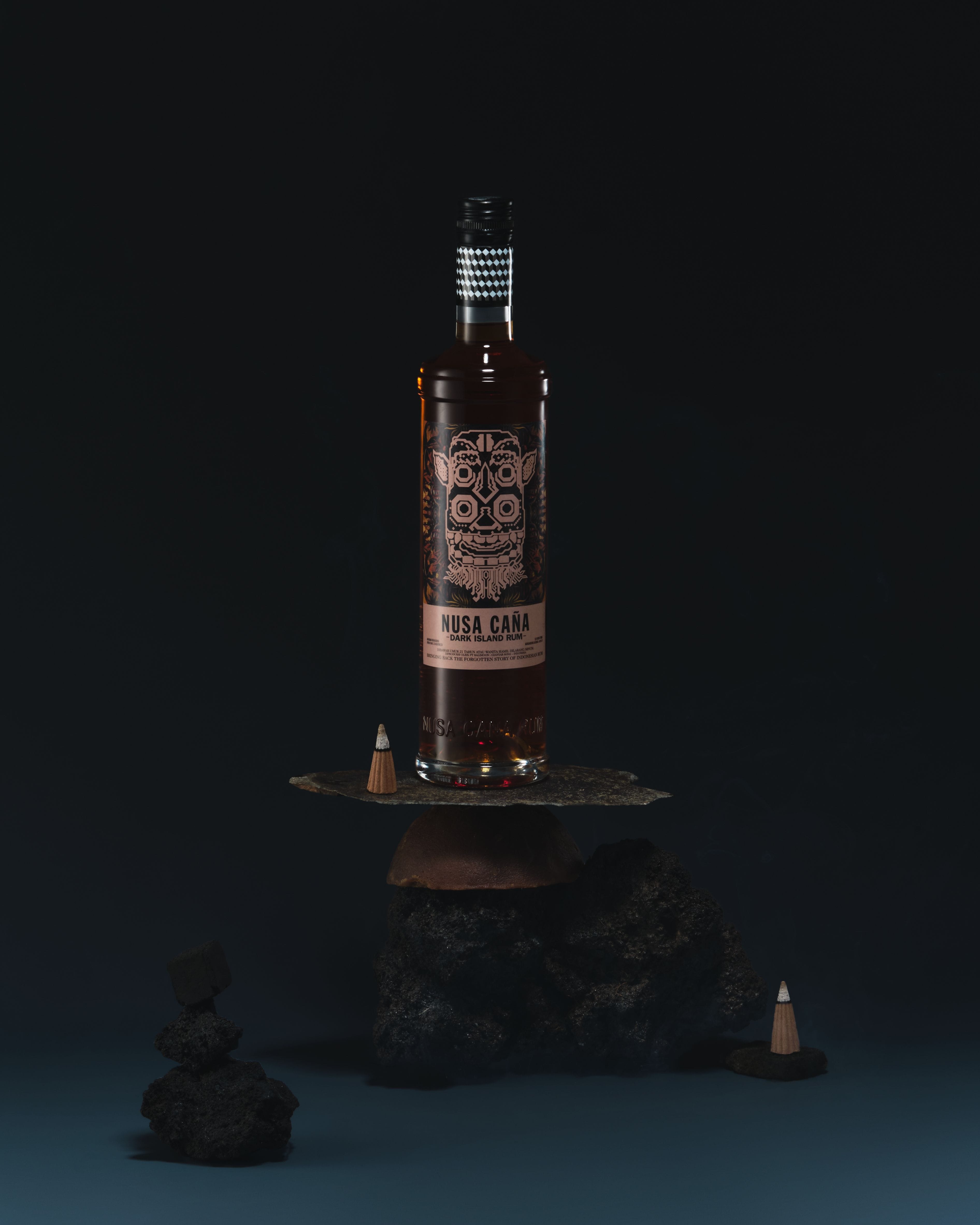 Nusa Caña Dark Island rum bottle with a dark sea-blue background placed on top lava rocks.